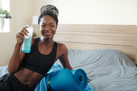 Portrait of smiling beautiful young Black woman with braided hair resting on bed and drinking...