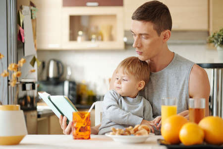Young father sitting at the table together with his little son and reading him a book during breakfastの写真素材