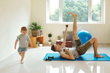 Young father lying on the floor on exercise mat and exercising with dumbbells while his son running around the roomの写真素材
