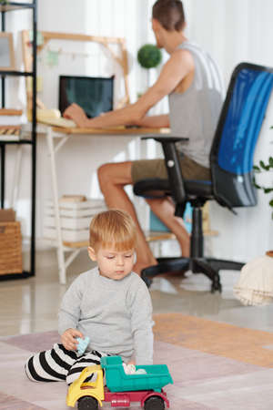 Child playing with toys with his father working on computer in the background at homeの写真素材