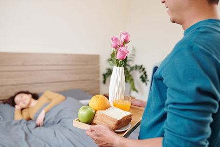 Cropped image of young man bringing tray with toasts, orange juice, fruits and flowers to surprise girlfriend sleeping in bedの写真素材