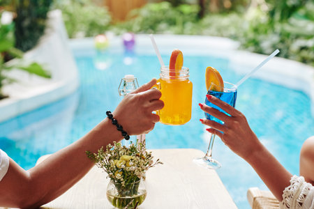 Hands of man and woman clinking glasses of sweet fruit cocktails when enjoying sunbathing by swimming poolの写真素材