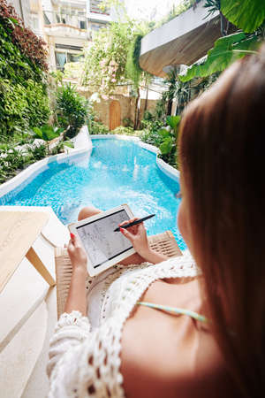Creative talented young woman relaxing by swimming pool and drawing fashion sketch on tablet computerの写真素材