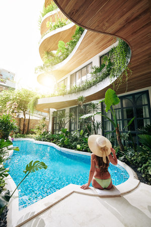 Young woman in bikini and straw hat resting by hotel swimming pool on sunny day with exotic plants...