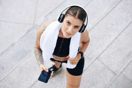 Pretty fit young woman with towel setting music in headphones via smartphone and looking up at cameraの写真素材
