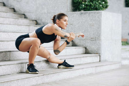 Serious fit young woman doing side lounge and stretching arm when exercising on steps after running on streetの写真素材