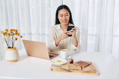 Pretty young Asian woman sitting at table, eating breakfast and checking messages on smartphone and e-mails on laptopの写真素材