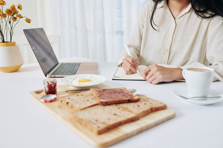 Businesswoman eating breakfast at home and writing in planner before having online meeting with colleaguesの写真素材