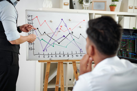 Young man attending lection on stock market trading and looking at broker pointing at whiteboad with...