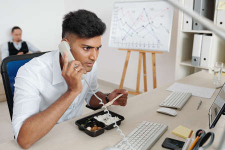 Stock market broker eating lunch at his office desk and answering phone call from clientの写真素材