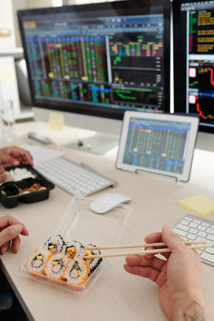 Cropped image of stock brokers eating sushi at office desk and discussing charts on computer...