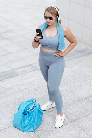 Pretty plus size fitness blogger standing outdoors after training and answering comments on social...