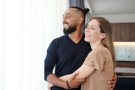 Portrait of happy embracing mixed-race couple hugging and looking through windowの写真素材