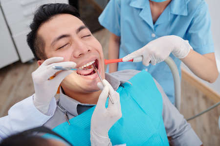 Dentist treating teeth of male patient when nurse putting saliva ejector inside mouthの写真素材