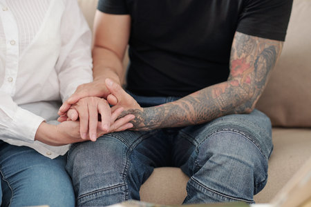 Hands of man with tattoed arms holding hands of his mature mother to reassure and support her