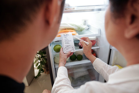 Senior woman checking her fridge, writing shopping list and asking adult son to buy her groceriesの写真素材