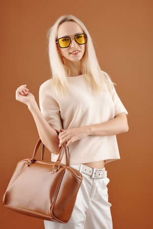 Portrait of stylish young woman in yellow glasses standing with leather bag and smiling at cameraの写真素材