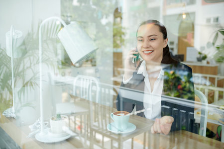 Happy young businesswoman drinking coffee in coffeeshop and talking on phone with friend or...