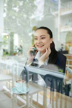 Cheerful young businesswoman talking on phone and drinking cup of coffee in coffeeshopの写真素材