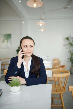 Portrait of young positive businesswoman sitting at cafe table and talking on phone with coworkerの写真素材