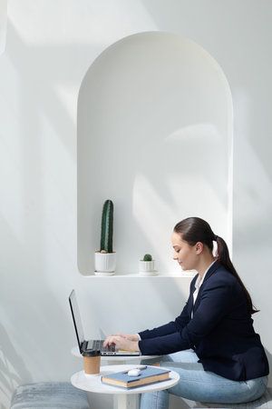 Smiling young female entrepreneur sitting at table in modern cafe and answering e-mails on laptop