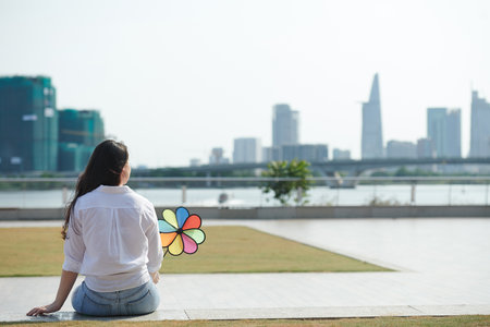 Young woman relaxing on river bank, holding pinwheel and looking at water, view from the backの写真素材