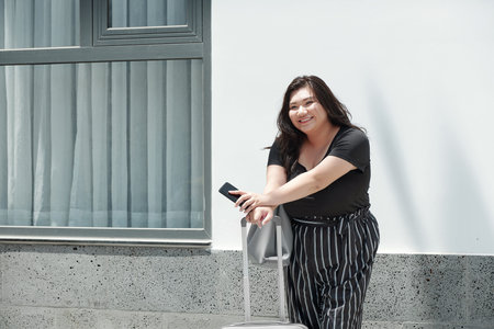 Cheerful pretty young blus size woman standing outdoors with suitcase and waiting for taxi car