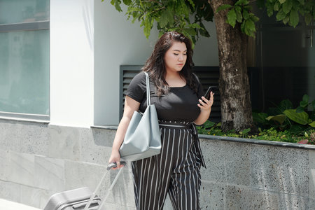 Pretty young serious woman with luggage walking on street and checking text messages on smartohone