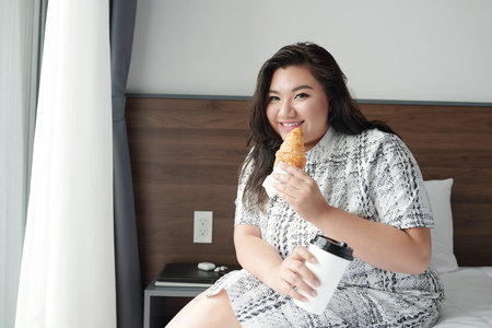 Portrait of smiling young woman sitting on bed and eating croissant with coffee for breakfast