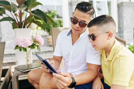 Father and son in sunglasses sitting in hotel backyard, reading information on tablet computer and...