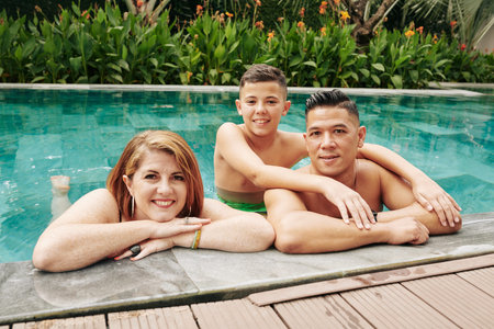 Joyful father, mother and kid standing in swimming pool and smiling at camera