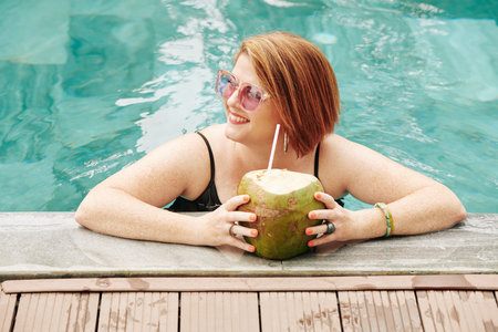 Happy mature woman with coconut cocktail standing in swimming pool and looking away