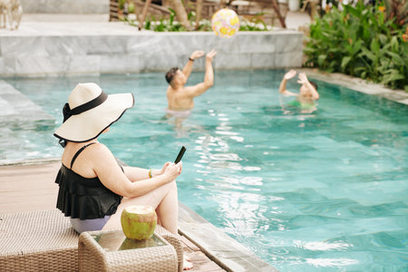 Woman sitting by swimming pool ans checking social media via smartphone when her husbad and son playing with inflatable ball in backgroundの写真素材