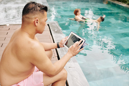 Serious fit man checking e-mails on tablet computer when his wife and soma playing in and splashing in swimming poolの写真素材