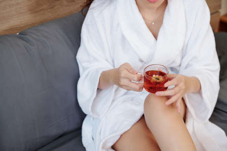 Cropped image of woman in bathrobe sitting on sofa with cup of tasty herbal teaの写真素材