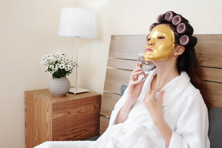 Young woman with golden rejuvenating face mask on massaging face with roller, spa day at home conceptの写真素材