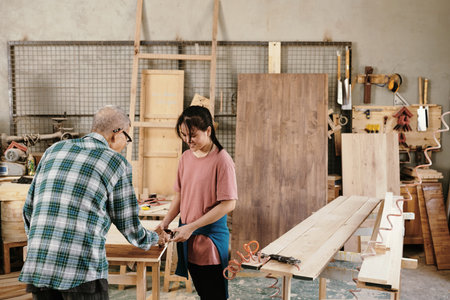 Senior Asian carpenter showing granddaughter how to cover sanded wood with oil, family business...