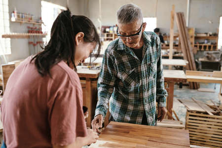 Experienced senior carpenter controlling his female apprentice applying oil or lacquer on wood boardの写真素材