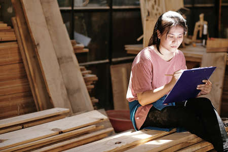 Creative young female customer sitting on pile of wooden boards and drawing furniture item blueprintの写真素材