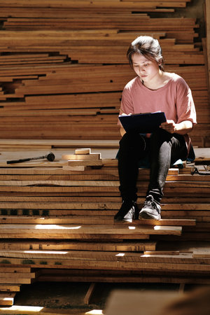 Young female carpenter working on design of furniture item for customer and drawing sketch she sitting on pile of wooden planksの写真素材