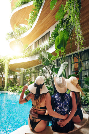 Happy young women in swimsuits and straw hats hugging and looking at beautful swimming pool of spa resort decorated with lush plantsの写真素材