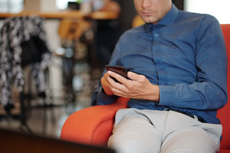 Cropped image of serious young entrepreneur sitting in armchair in coffeeshop and answering text...