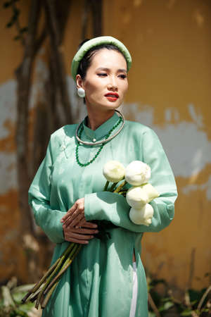 Portrait of beautiful young Vietnamese woman in traditional dress and headwer holding lotus flowers and looking awayの写真素材