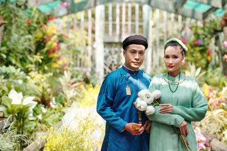 Just married Asain man and woman in ao dai dresses and headwear called khan dong standing in greenhouse with beautiful blooming flowersの写真素材