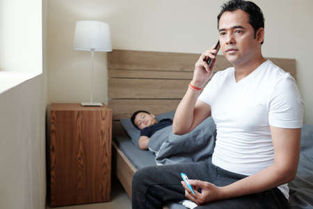 Worried Indian man sitting on bed of his sick child with fever and calling to hospitalの写真素材