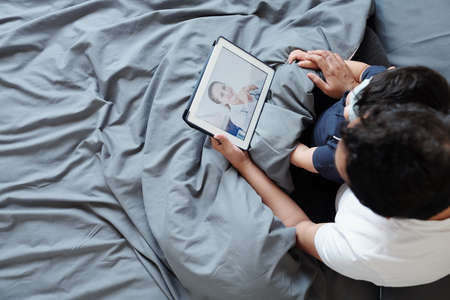 Father hugging his kid when they are sitting in bed and video calling doctor to discuss flu treatmentの写真素材