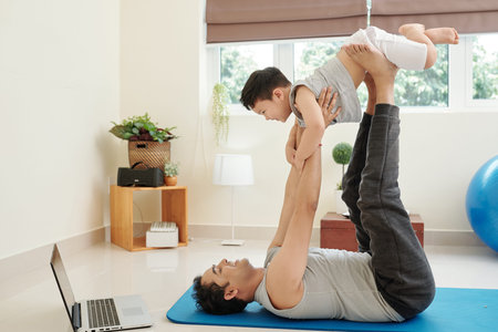 Smiling Indian man lying on yoga mat and playing with his laughing little son