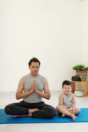 Laughing little boy sitting next to his father trying to meditate at homeの写真素材