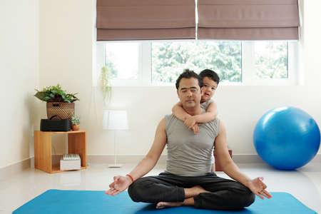 Little child hugging his father meditating at home in lotus position trying to find inner pieceの写真素材