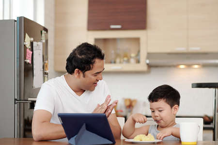 Indian man clapping to his son eating plate of mashed potato by himself at kitchen tableの写真素材